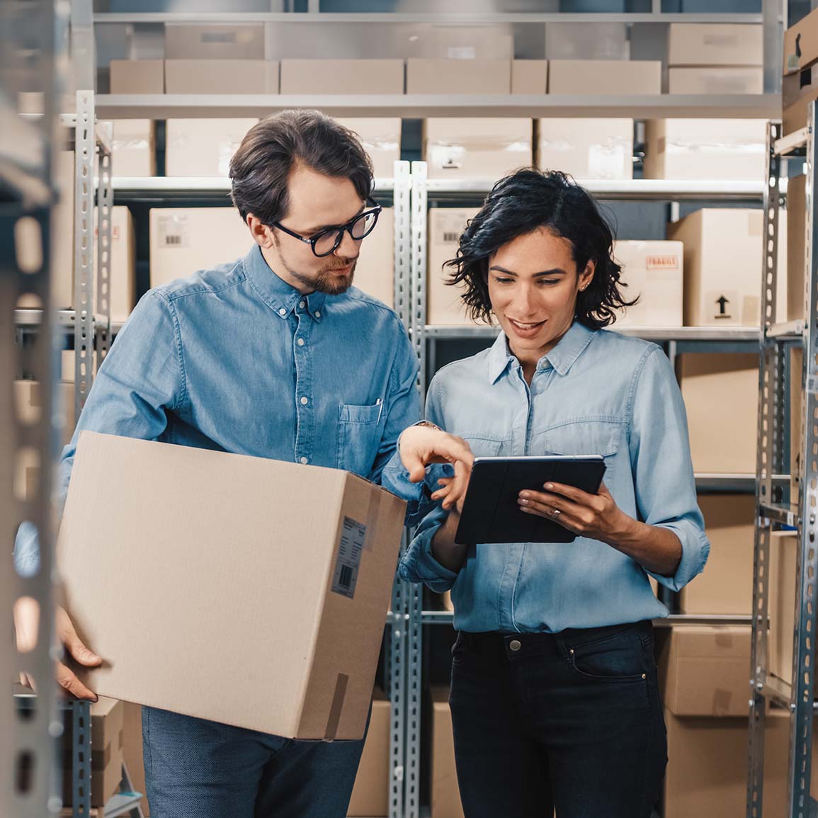 two employees in a warehouse holding a package and looking at a tablet